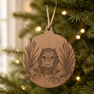May include: Wooden Christmas ornament featuring a detailed engraving of a swamp creature emerging from water, surrounded by reeds. The ornament is round with a decorative top and is suspended by a natural twine string. The design is in shades of brown and black.