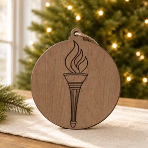 May include: A round, wooden ornament with a torch design. The torch is etched into the wood and features flames at the top. A jute string is attached for hanging. The ornament is a light brown color.