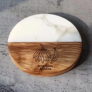 May include: Round coaster with a two-tone design. The top half is white with a marble-like pattern, and the bottom half is wood-grained. The wood section features a banana illustration and the text "BLUE JAVA BANANA".