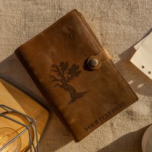 May include: A brown leather journal with a tree design and the text "YOUR TEXT HERE". The journal has a button closure and is lying on a textured surface. A wooden cutting board and a light bulb are in the background.