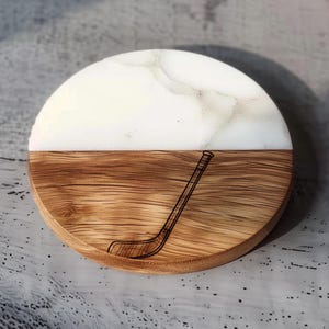 May include: A round coaster with a hockey stick design. The top half is white with a marble effect, and the bottom half is wood-grained. The hockey stick is etched into the wood. A decorative item for home or office.