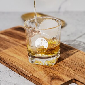 May include: A clear glass with a rocket ship design and the text "Your Custom Text Here" is filled with a golden liquid and an ice cube. The glass is sitting on a wooden cutting board.