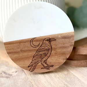May include: A round wooden coaster with a white top half and a brown bottom half. A detailed engraving of a raven with a crescent moon above it is on the brown section. The coaster is on a wooden surface.