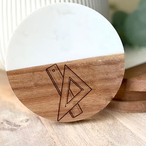 May include: A round coaster with a white top and a wooden bottom. The wooden section features an engraved design of a ruler and a triangle. The coaster is a decorative item.
