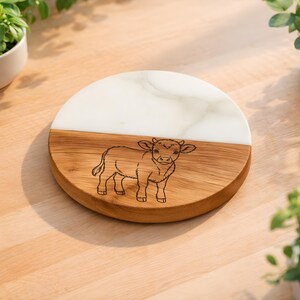 May include: A round wooden and white marble coaster with a laser-etched illustration of a calf. The coaster is split diagonally, with the top half in white marble and the bottom half in wood. The calf is centered on the wooden portion.