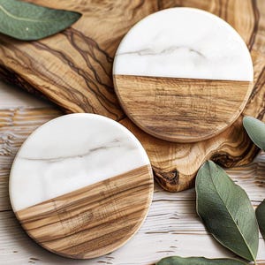 May include: Two round coasters with a half-and-half design. The top half is white marble, and the bottom half is wood. The coasters are on a wooden surface with green leaves. The coasters are approximately 4 inches in diameter.