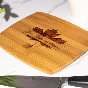 May include: A rectangular bamboo cutting board with a maple leaf design and the text "Your Custom Name" engraved on it.