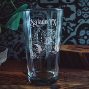 May include: A clear glass pint glass featuring a white etched illustration of a historic mill with a water wheel, a stone bridge, and trees. The text Salado, TX is printed in a white serif font above the detailed landscape scene on the glass.