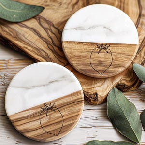 May include: Two round coasters with a half-and-half design. The top half is white marble, and the bottom half is wood. A black outline of a tomato is etched on the wood. The coasters are on a wooden cutting board.