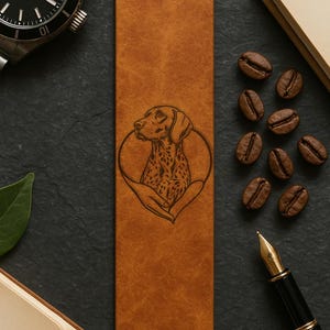 May include: A brown leather bookmark featuring a detailed engraved image of a dog within a heart shape. The bookmark is rectangular and lies on a dark surface, accompanied by coffee beans, a pen, a watch, and a book.