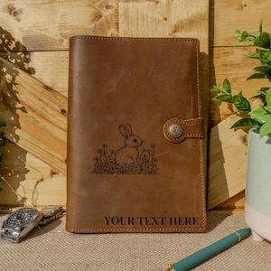May include: Brown leather journal with a laser engraved design of a rabbit sitting in a field of flowers. The text "YOUR TEXT HERE" is engraved below the design.