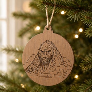 May include: A round wooden Christmas ornament with a Bigfoot design. The ornament features a detailed engraving of the legendary creature emerging from water or mist. It is suspended by a natural twine string, ready for display on a Christmas tree.