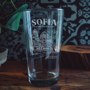 May include: A clear glass pint tumbler featuring a white etched design of the Alexander Nevsky Cathedral in Sofia, Bulgaria. The text SOFIA and BULGARIA is printed above the architectural illustration, which includes a lion statue on a pedestal.