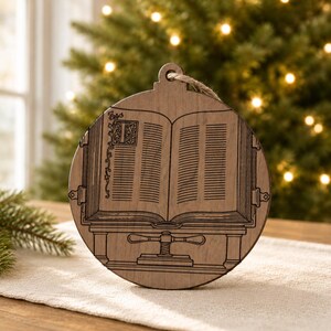 May include: A round wooden Christmas ornament with a detailed engraving of an open book on a stand. The ornament has a natural wood finish and a jute twine loop for hanging. The book's pages and decorative elements are intricately etched.