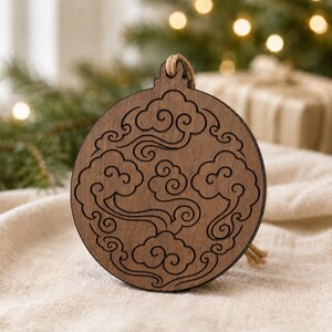 May include: A round, wooden Christmas ornament with a cloud design. The ornament is brown with a darker brown cloud pattern. It has a jute string for hanging. The background includes a Christmas tree and a gift.