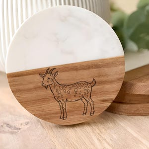 May include: A round wooden coaster with a white marble-look top and a goat illustration. The bottom half is natural wood. The goat is brown with a spotted pattern. The coaster is next to a wooden serving board.