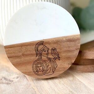 May include: Round wooden coaster with a white top half and a brown bottom half. The brown section features a laser-etched design of a figure in a helmet with an owl and shield. The coaster is likely for home decor.