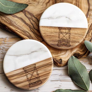 May include: Two round coasters with a white marble top and a wooden base. The wooden base features an engraved hourglass design. The coasters are displayed on a wooden cutting board with green leaves.