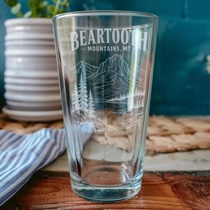 May include: A clear glass pint glass featuring a white etched design of the Beartooth Mountains, MT. The graphic includes a detailed mountain range, evergreen trees, and a body of water, with the text Beartooth Mountains, MT printed above.