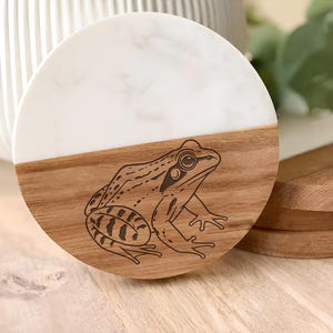 May include: A circular coaster featuring a split design with a white marble top half and a natural wood bottom half. The wooden section is engraved with a detailed black line drawing of a frog in a seated position, showcasing its textured skin.