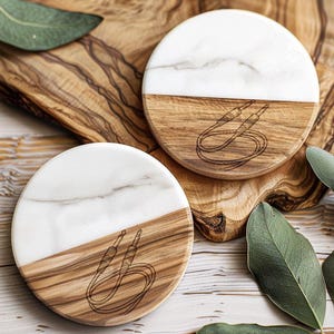 May include: Two round coasters with a half-and-half design. The top half is white marble, and the bottom half is wood-grain. A black line drawing of a cable with two jacks is etched into the wood. The coasters are on a wooden surface.