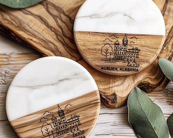 May include: Two round coasters with a half-and-half design. The top half is white marble, and the bottom half is wood. Each coaster features a black line drawing of a building, tree, and tiger, with the text "AUBURN, ALABAMA" below.