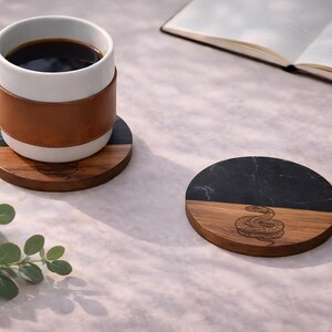 a white mug with a brown leather sleeve, a wooden coaster with a black marble top, and an open book on a table.