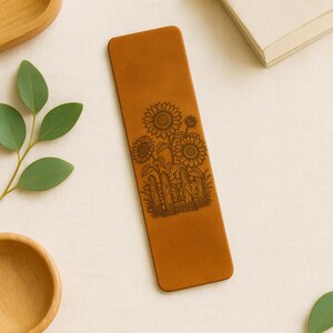 May include: A rectangular, tan leather bookmark featuring a detailed laser-etched design of sunflowers and a wooden fence. The bookmark is a warm brown color and has rounded corners. The sunflowers are in full bloom.
