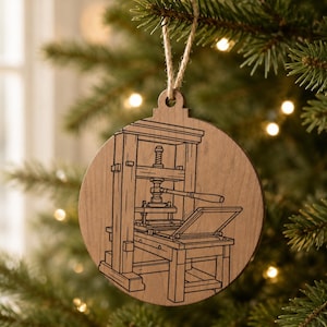 May include: Wooden Christmas ornament featuring a detailed line drawing of a printing press. The ornament is round with a cutout at the top for hanging. The design is etched into the wood, showcasing the press's intricate details.