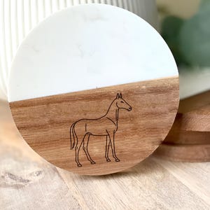 May include: A round coaster with a white and wood grain design. A black outline of a horse is etched into the wood grain section. The coaster is made of wood and resin.