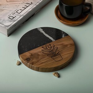 May include: A round coaster with a half-and-half design. One half is black marble with white veining, and the other is light brown wood. A potted plant is etched into the wood. A black mug sits on a wooden coaster.