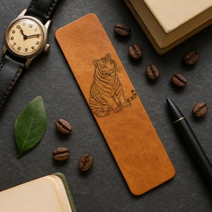 May include: A tan leather bookmark with a detailed engraved tiger design. The bookmark is rectangular and lies on a dark surface, surrounded by coffee beans, a watch, a pen, a leaf, and a book.