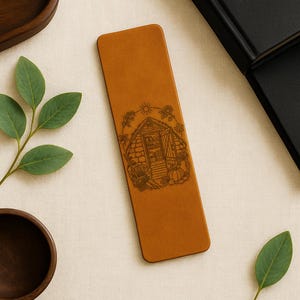 May include: A tan leather bookmark with a detailed engraved illustration of a garden shed, sun, and sunflowers. The bookmark is rectangular and has rounded corners. The design includes pumpkins and other garden elements.