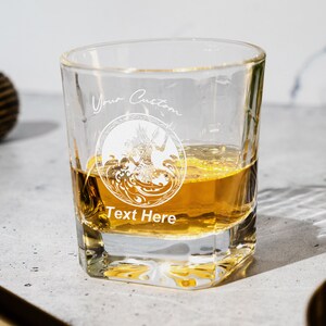 May include: A clear glass rocks glass with a square base filled with amber liquid. The glass has a white design etched into the side with the text "Your Custom Text Here".