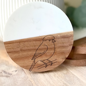 May include: A round coaster with a bird design. The top half is white, and the bottom half is wood-toned. A black outline of a bird perched on a branch is etched into the wood. The coaster is on a wooden surface.