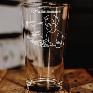May include: Clear pint glass with a white etched design. The design features a cartoon software engineer at a computer with the text "SOFTWARE ENGINEER" above. A mug is next to the engineer. The glass is on a wooden surface.