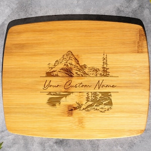 May include: A rectangular bamboo cutting board with a laser engraved design of a mountain range reflected in a lake. The text "Your Custom Name" is engraved below the image.