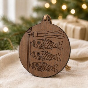 May include: Wooden ornament with a brown finish, featuring an engraved design of three carp streamers and a banner. The ornament has a jute string for hanging and is round with a flat back.
