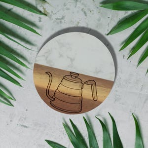A white circular plate with a black and white illustration of a coffee pot is surrounded by green leaves on a white background.