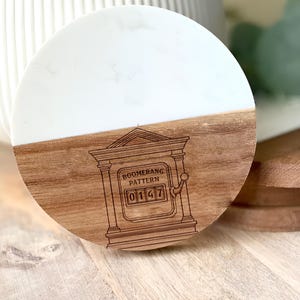 May include: A round wooden and white marble coaster with the text "BOOMERANG PATTERN" and the numbers "0147" etched into the wood. The coaster is half wood and half white marble.