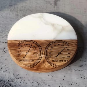 May include: A round coaster with a white marble top and a wooden bottom. The wooden section features a barometer and hygrometer design. The barometer shows "Fair" and "Change" with weather symbols. The hygrometer shows humidity levels.