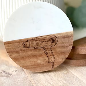 May include: A round wooden coaster with a white marble-like top. The bottom half is natural wood, engraved with a heat gun design and the words "HEAT GUN". A decorative item for a workshop or craft space.