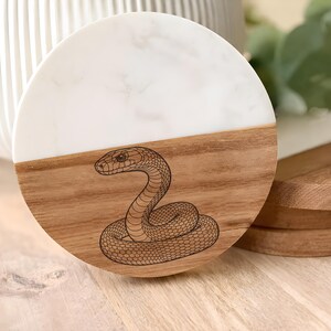 May include: A round coaster with a two-tone design. The top half is white with a marble pattern, and the bottom half is wood. A detailed illustration of a snake is etched into the wood. Additional wooden coasters are visible.