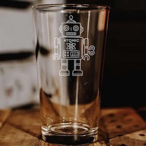 May include: A clear pint glass with a white etched robot design. The robot has a head, eyes, and a body with the word "ATOMIC" and a symbol. The glass is on a wooden surface.