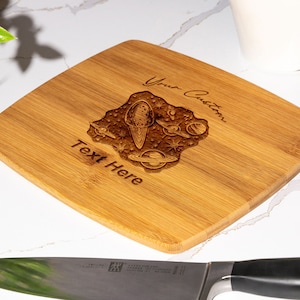 May include: A light brown rectangular bamboo cutting board with a laser engraved design of a rocket ship, planets, and stars. The text "Your Custom Text Here" is engraved on the board.