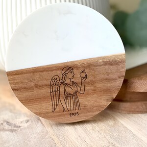 May include: A round coaster with a white and wood grain design. The wood grain section features a laser-etched illustration of a winged figure holding an apple, with the name "ERIS" below. The coaster is likely made of wood and resin.