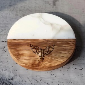 May include: A round coaster with a two-tone design. The top half is white with a marbled pattern, and the bottom half is wood-grained with a carved lyre and wing design. The coaster is made of wood and resin.