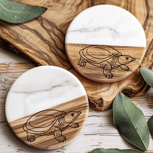 May include: Two round coasters with a turtle design. Each coaster is split diagonally, with a white marble section and a wooden section featuring a turtle illustration. The coasters are on a wooden surface, with green leaves scattered around.