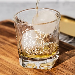 May include: A clear, textured glass with a white etched dog face design, filled with amber liquid and a large ice sphere. The glass sits on a wooden cutting board, with a gold tray in the background. A stream of liquid is being poured into the glass.