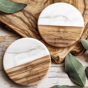 May include: Two round coasters with a half-and-half design. The top half is white marble, and the bottom half is wood with a sun and table engraving. The coasters are on a wooden cutting board with green leaves.